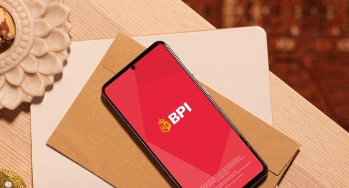 A smartphone displaying the BPI mobile banking app on a wooden table, accompanied by a brown envelope and a decorative item.
