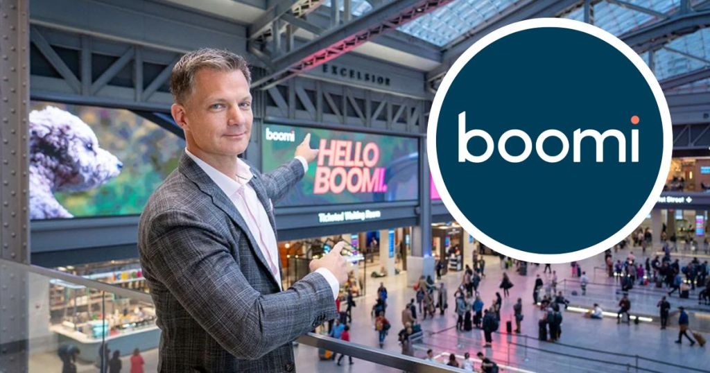 A person in a blazer gestures towards a large digital display that says 'HELLO BOOMI' in a busy indoor space with people walking around.