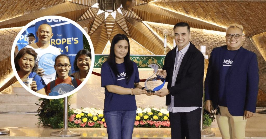 A group of individuals celebrate a choral competition, with a woman in a blue shirt receiving an award from a man in a black jacket. Behind them, floral decorations are visible in a church setting.
