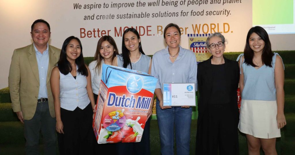 A group of seven individuals standing together at an event, holding a Dutch Mill product and an award certificate, with a backdrop displaying a sustainability message.