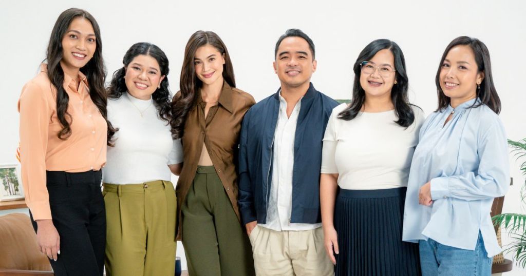 Group of six professionals posing together, dressed in business casual attire, smiling at the camera in a well-lit indoor setting with light-colored background.