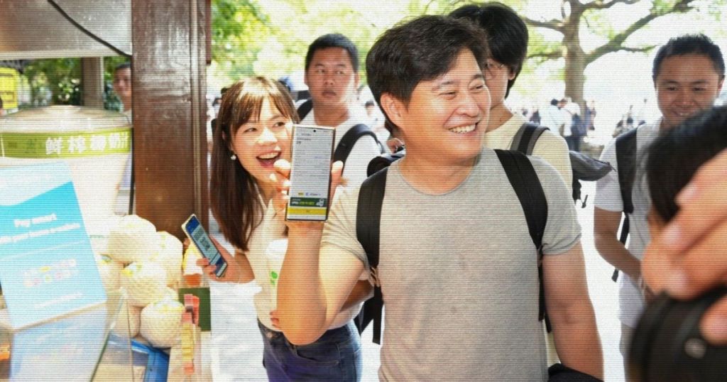 A group of individuals at an outdoor food stall, with one person smiling and holding up a smartphone displaying a payment app, while others are engaged and looking towards the stall.
