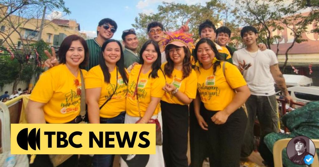 A group of smiling individuals wearing bright yellow shirts, standing together for a photo outdoors, with trees and buildings in the background. Some members are making peace signs and displaying joyful expressions, suggesting a festive atmosphere.