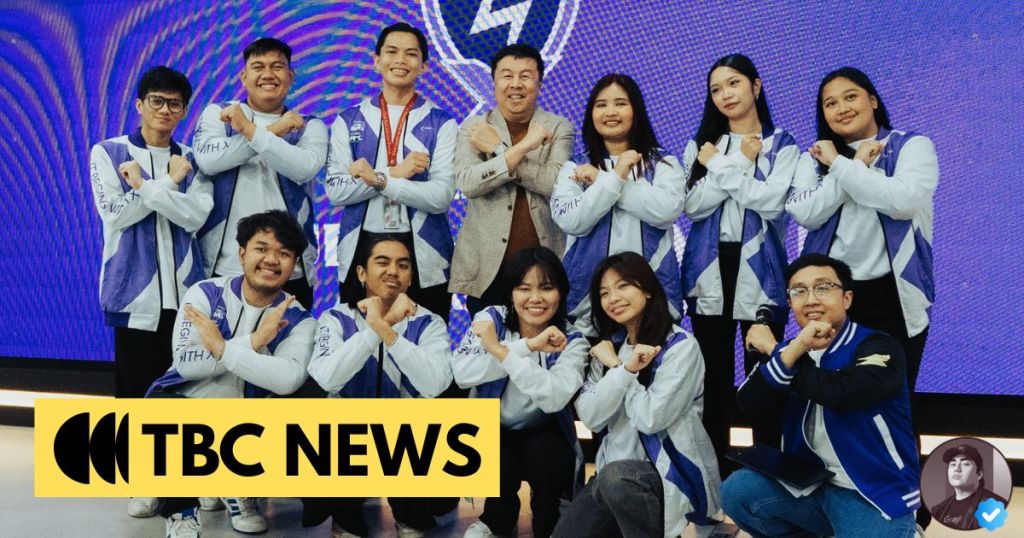 A group of eleven individuals posing together for a photo, all wearing matching jackets with blue and white colors, smiling and gesturing in a creative manner. The background features a vibrant digital display and the TBC News logo is prominently displayed at the bottom.