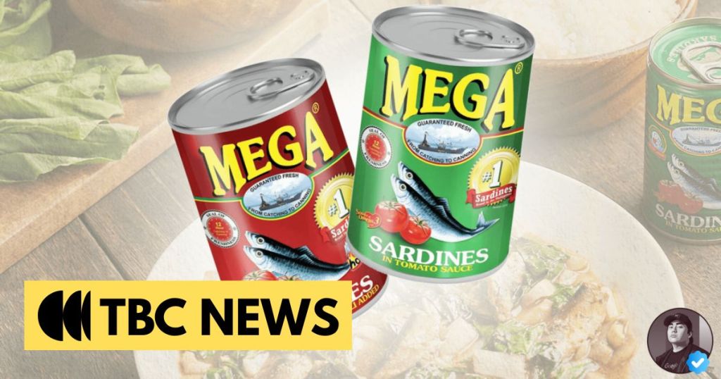Two cans of Mega brand sardines in tomato sauce, one red and one green, displayed against a blurred background of a wooden table and a bowl of food. TBC News logo in the foreground.