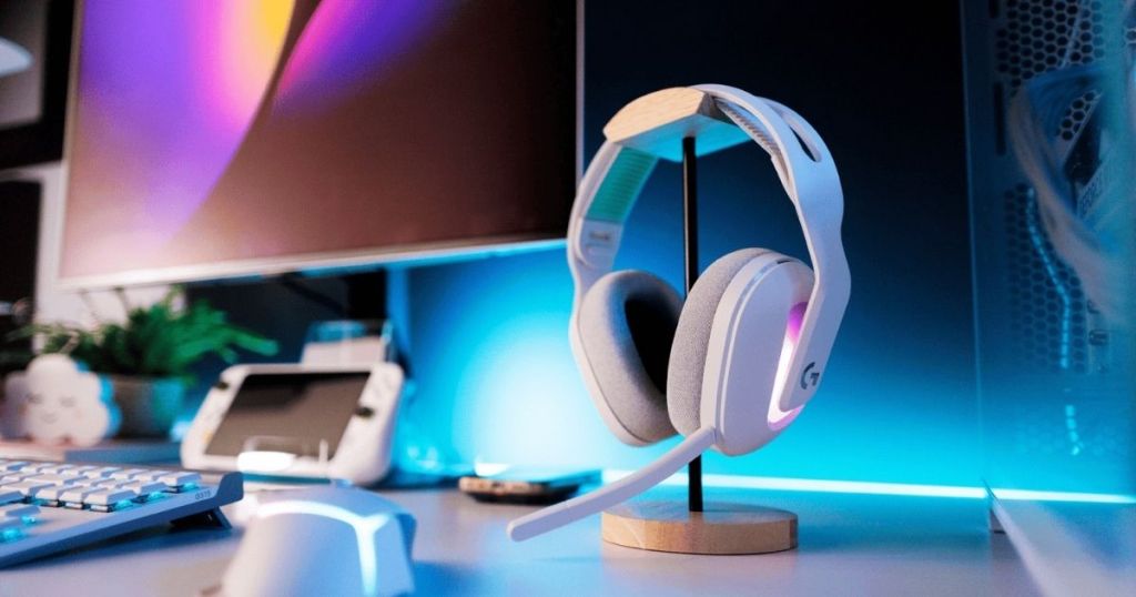 A white gaming headset displayed on a wooden stand, featuring gray ear cushions and RGB lighting, with a computer monitor in the background and a keyboard and mouse on the desk.