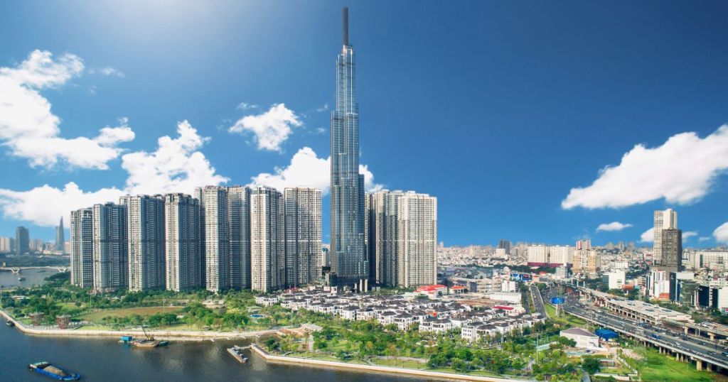 A panoramic view of a modern city skyline featuring a tall skyscraper surrounded by high-rise buildings, greenery, and a river under a clear blue sky.
