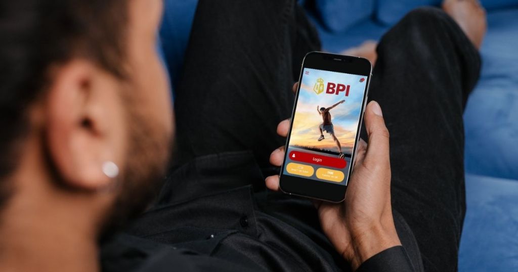 A person sitting on a couch, holding a smartphone displaying the BPI banking app login screen against a colorful sunset background.