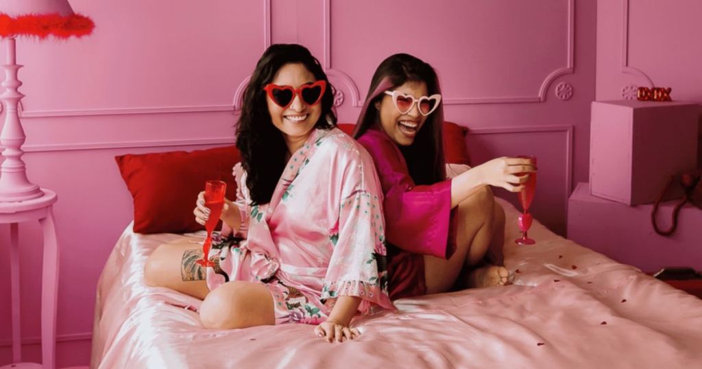 Two women sitting on a bed in a pink room, wearing colorful robes and heart-shaped sunglasses, each holding a glass of a pink drink, with a playful and cheerful expression.