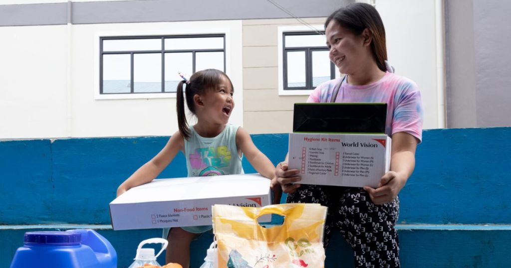 A young girl and a woman smiling at each other while holding boxes of hygiene kits and non-food items, with various bags and containers nearby.