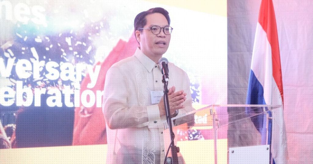 A speaker in traditional attire delivering a speech at an anniversary celebration event, with a backdrop displaying celebratory graphics and a flag in the scene.