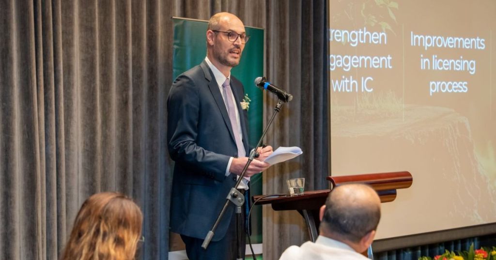 A man in a suit speaks at a podium during an event, with a presentation backdrop displaying key points about engagement and licensing improvements.
