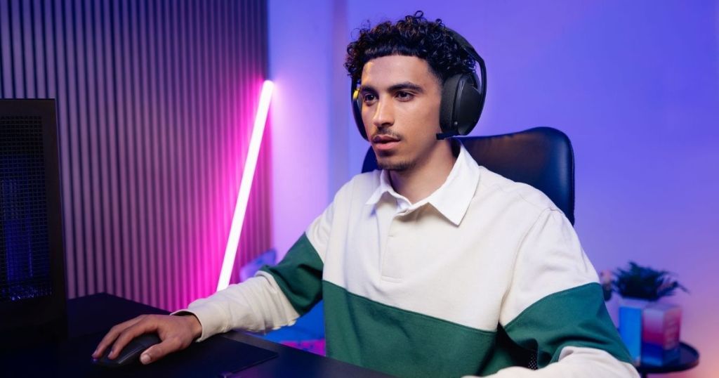 A young man wearing a headset and a color-blocked sweater sits at a gaming desk, focused on a computer screen, with colorful LED lights in the background.