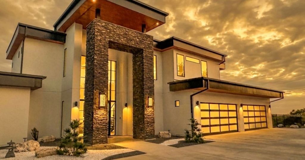 Modern house exterior with stone accents, large windows, and a two-car garage, set against a dramatic sunset sky.