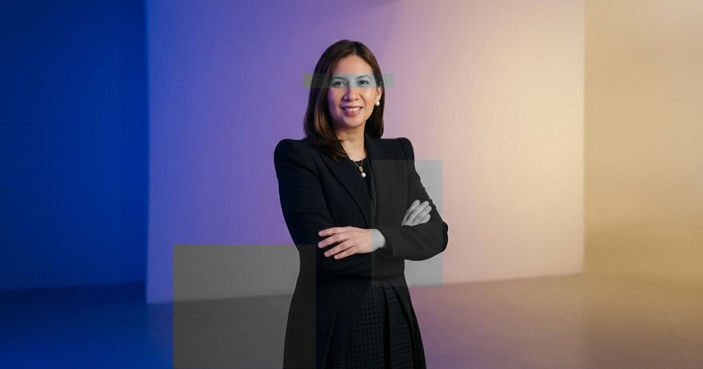 A confident woman in a black blazer stands with her arms crossed against a softly lit background featuring blue and beige hues.