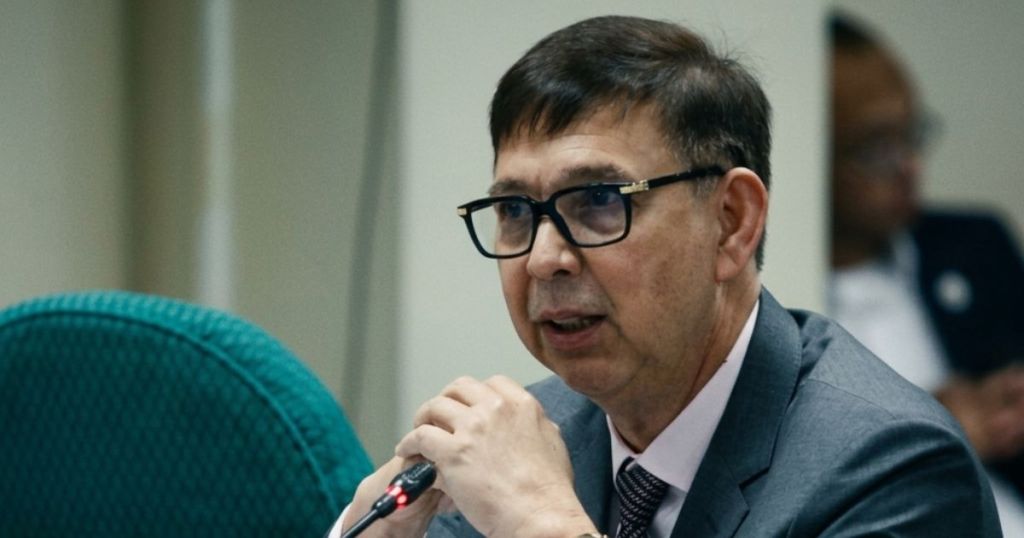 A middle-aged man with glasses speaks into a microphone during a meeting, wearing a suit and tie.