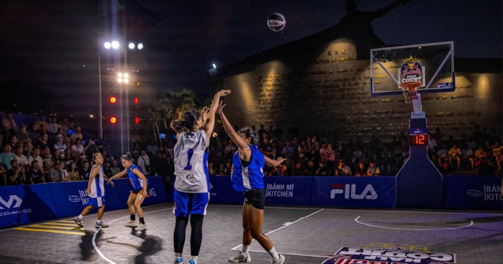 A basketball game in progress at night, with players in blue and gray jerseys shooting and defending on an outdoor court, surrounded by a cheering crowd under bright lights.