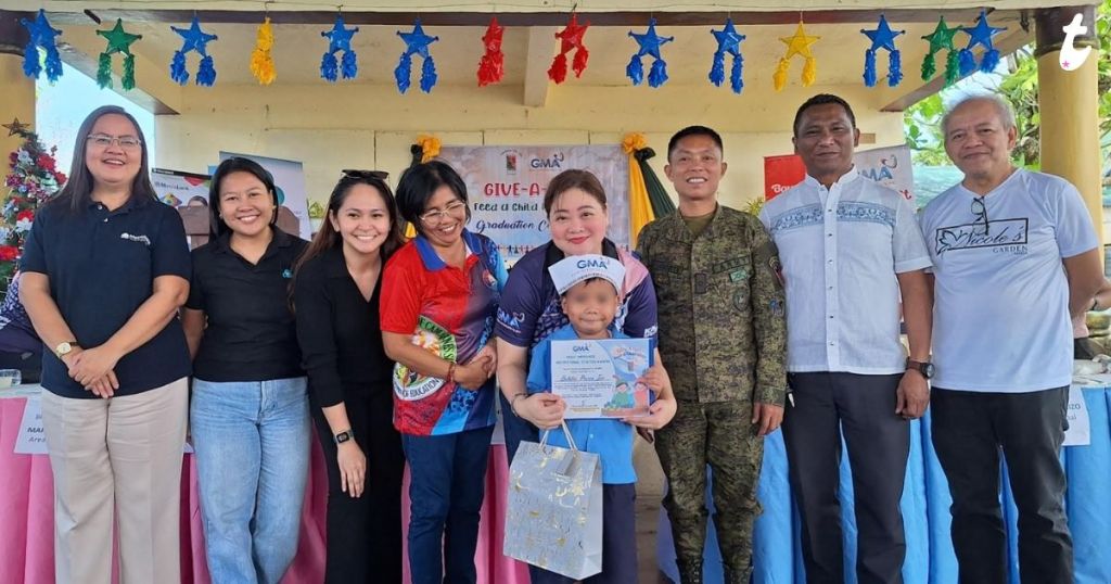 A group of individuals poses together at an event, with decorations and a banner in the background. The scene includes a woman holding a child and a certificate, a soldier in uniform, and several others smiling at the camera.