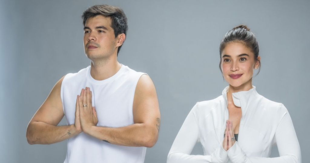 A man and woman standing side by side in a prayer pose, wearing athletic clothing. The man is in a sleeveless white shirt, and the woman is in a fitted white jacket. They are both smiling, with a gray background.