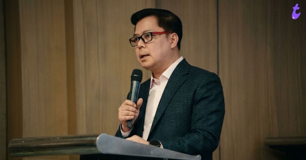 A man in a dark blazer and glasses speaking into a microphone while standing at a podium in a conference setting.