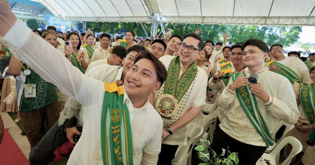 A group of graduates in formal attire celebrate at a ceremony, posing for a selfie with smiles and excitement, while others cheer in the background.