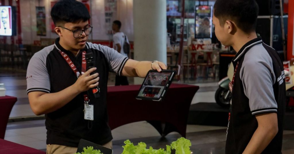 A person presenting information on a tablet to another individual in a public space, with a table featuring lettuce in the foreground.