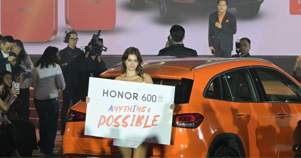 A model holding a sign that reads 'HONOR 600 5G series ANYTHING'S POSSIBLE' at a product launch event, in front of an orange car with a presentation backdrop and an audience.