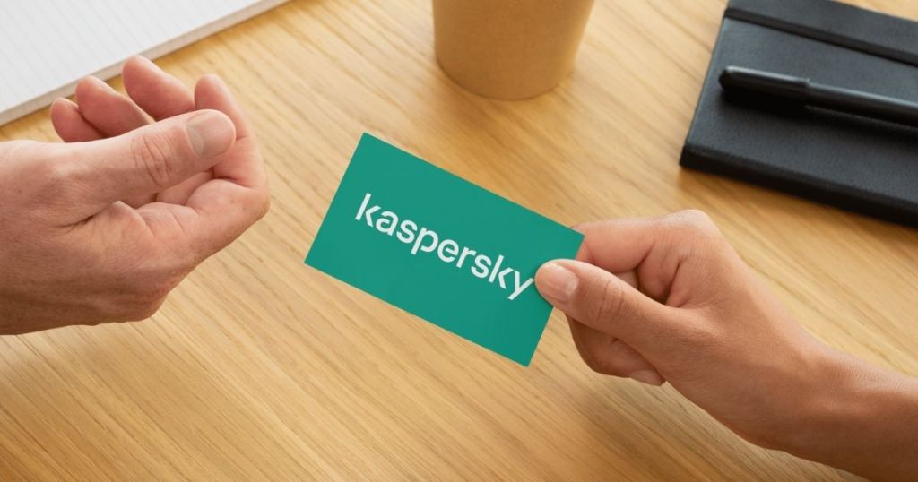 A business card with the Kaspersky logo being exchanged between two hands on a wooden table, with a coffee cup and a notepad in the background.