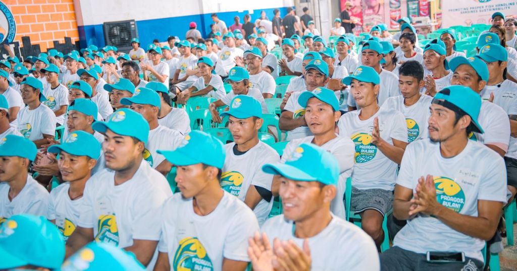 A large group of men wearing matching white t-shirts and turquoise caps, seated in a hall with green chairs, attentively clapping and engaging during an event.