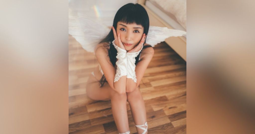 A young woman with angel wings and elegant gloves, sitting on a wooden floor, posing with a playful expression.