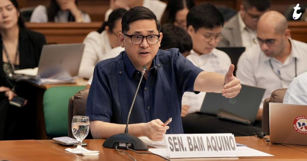 A man wearing glasses and a dark blue shirt gestures while speaking during a meeting, with a nameplate indicating he is Senator Bam Aquino. In the background, several people are seated with laptops.