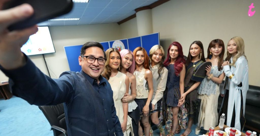 A group of eight people, including a man taking a selfie, are posing together in a room. They are smiling and wearing fashionable outfits, with a blue backdrop behind them.