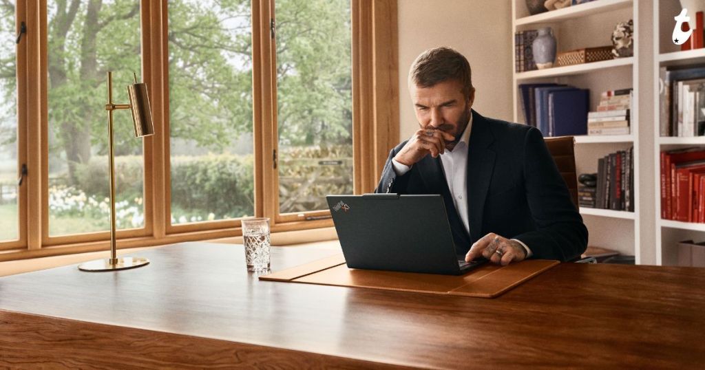 A man in a suit sits at a wooden desk, thoughtfully looking at his laptop in a well-lit room with large windows overlooking a garden.