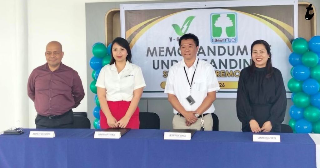 A group of four individuals standing in front of a banner that reads 'Memorandum of Understanding Signing Ceremony'. They are positioned behind a table with nameplates and decorations featuring green and blue balloons.
