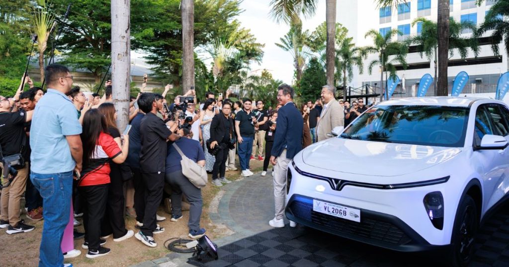 A large crowd of people, including media and fans, gathers around a white electric car, with two men in front of it at an outdoor event.