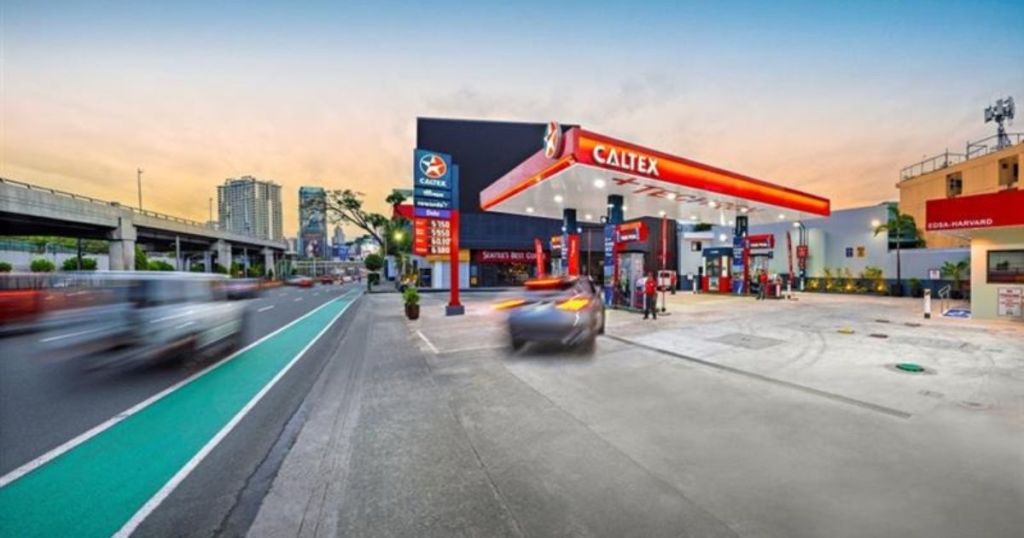 A Caltex gas station at sunset, with multiple fuel pumps, a price display, and vehicles passing on a nearby road.