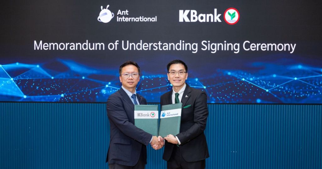 A signing ceremony for a Memorandum of Understanding between Ant International and KBank, featuring two men holding documents in front of a digital display.