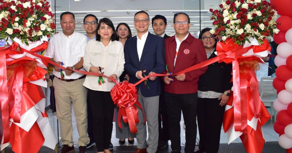 A group of people gathered for a ribbon-cutting ceremony, featuring large floral arrangements and red ribbons. They are smiling and holding scissors, preparing to cut the ribbon to mark the opening of an event or establishment.