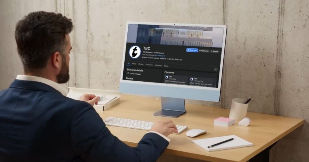 A person in a business suit sitting at a desk, looking at a computer screen displaying a social media profile for TBC. The workspace has a modern aesthetic with a cup and stationery on the table.