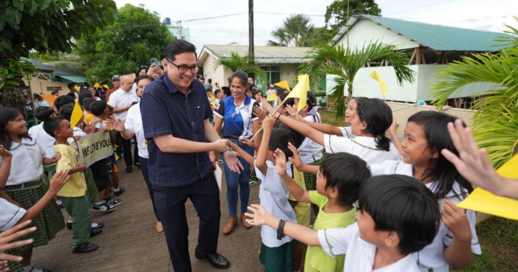 A group of children excitedly greet a smiling man as he walks down a path, surrounded by greenery and supportive adults, holding yellow flags and signs.
