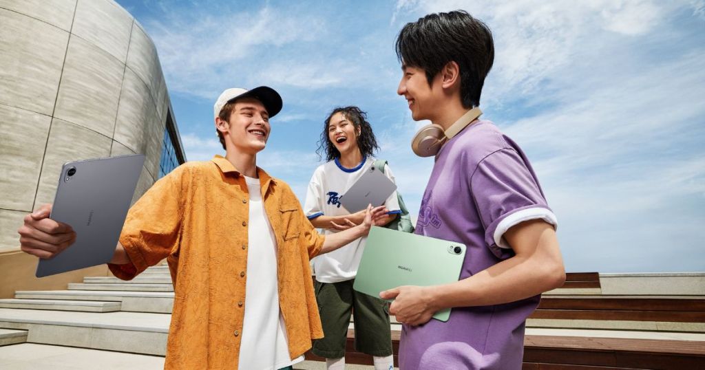 A group of three young people outdoors, smiling and interacting while holding tablets. One person wears an orange shirt and holds a gray tablet, another is in a white shirt and blue jacket, and the third in a purple shirt is holding a green tablet. They are surrounded by modern architecture and a blue sky.