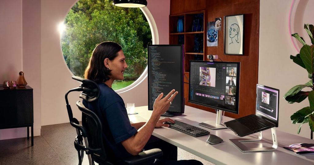 A person sitting at a modern desk with multiple computer screens, engaged in a video call, in a stylish workspace with a round window and a view of greenery.
