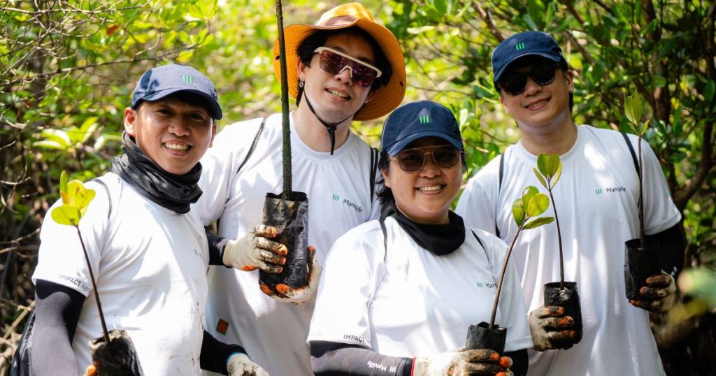 Four individuals smiling and holding young plants in pots while standing in a lush mangrove environment, wearing matching white shirts and caps.